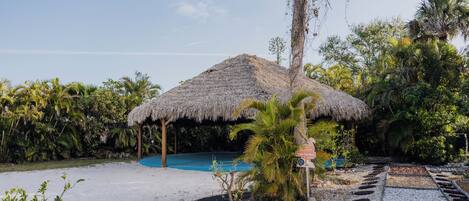 A thatched-roof structure with a pool, surrounded by tropical vegetation and a pebble path.
