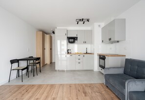 Eine moderne Küche mit weißen Schränken, einer Mikrowelle und einem Herd. Es gibt einen Esstisch mit Stühlen und ein graues Sofa.