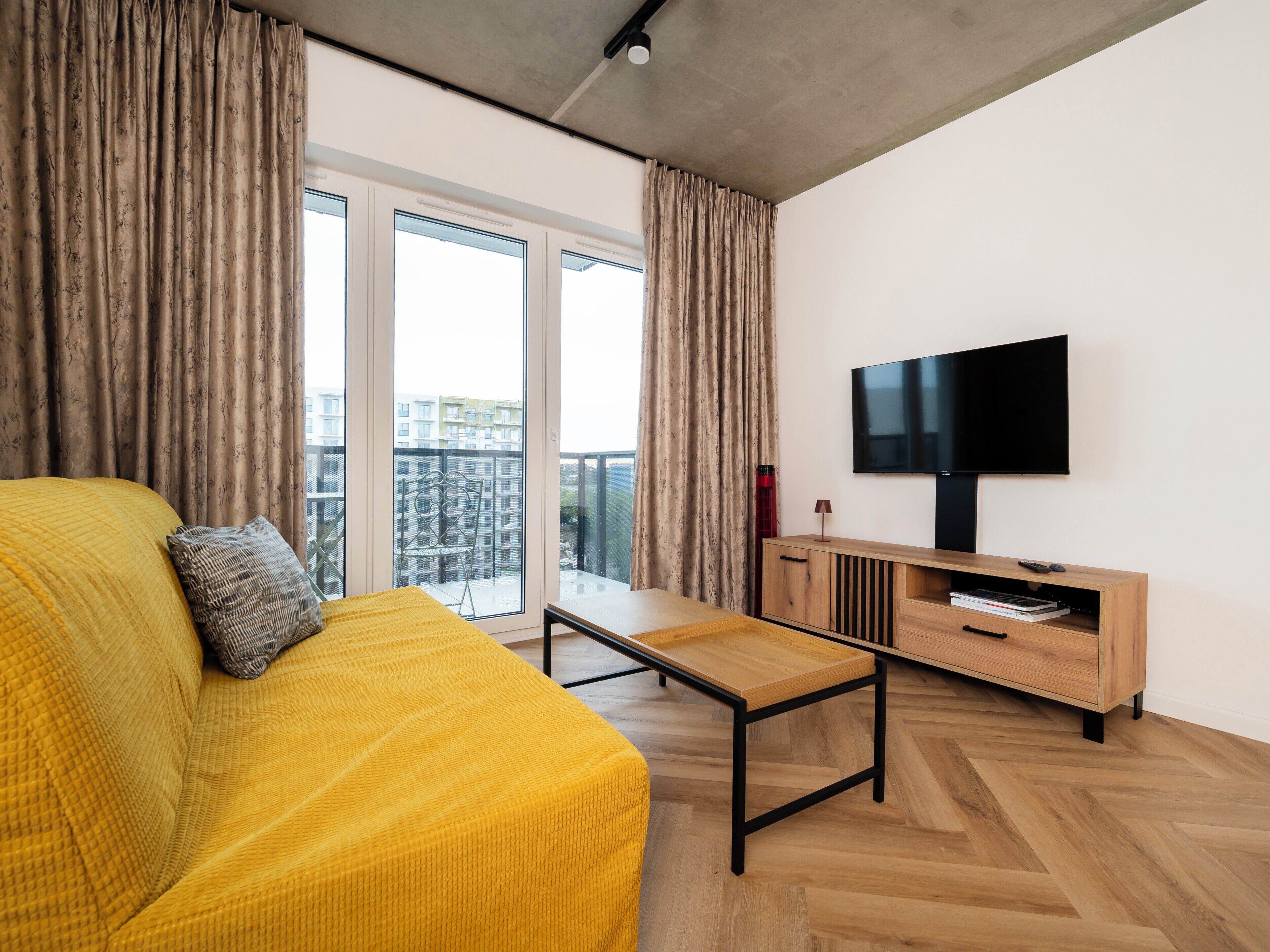 Un salon moderne avec un canapé jaune, une table basse en bois, un téléviseur posé sur un meuble en bois et une grande fenêtre avec des rideaux.