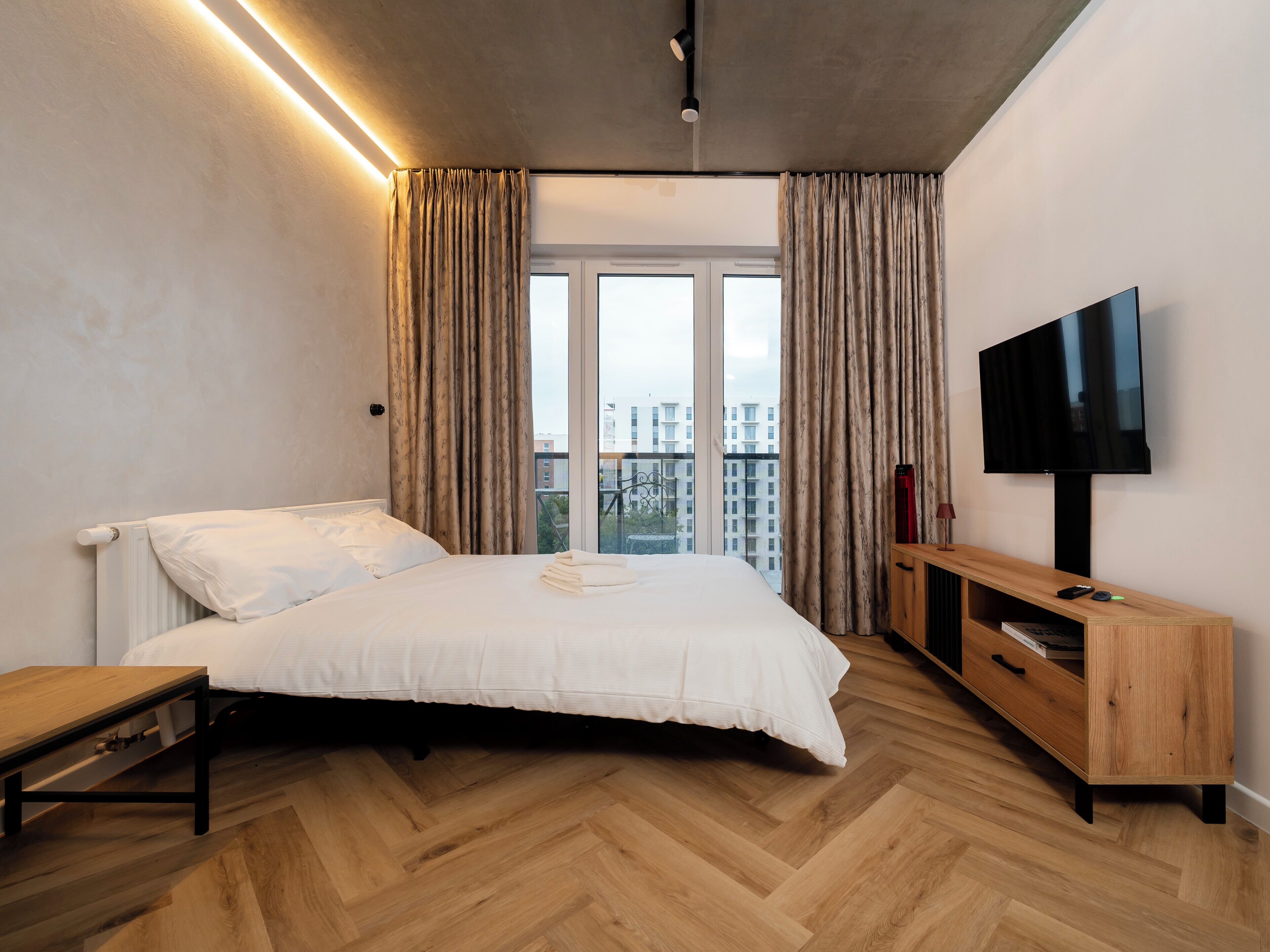 Une chambre moderne avec un lit, un plancher en bois, des rideaux, un téléviseur et une vue sur les bâtiments.