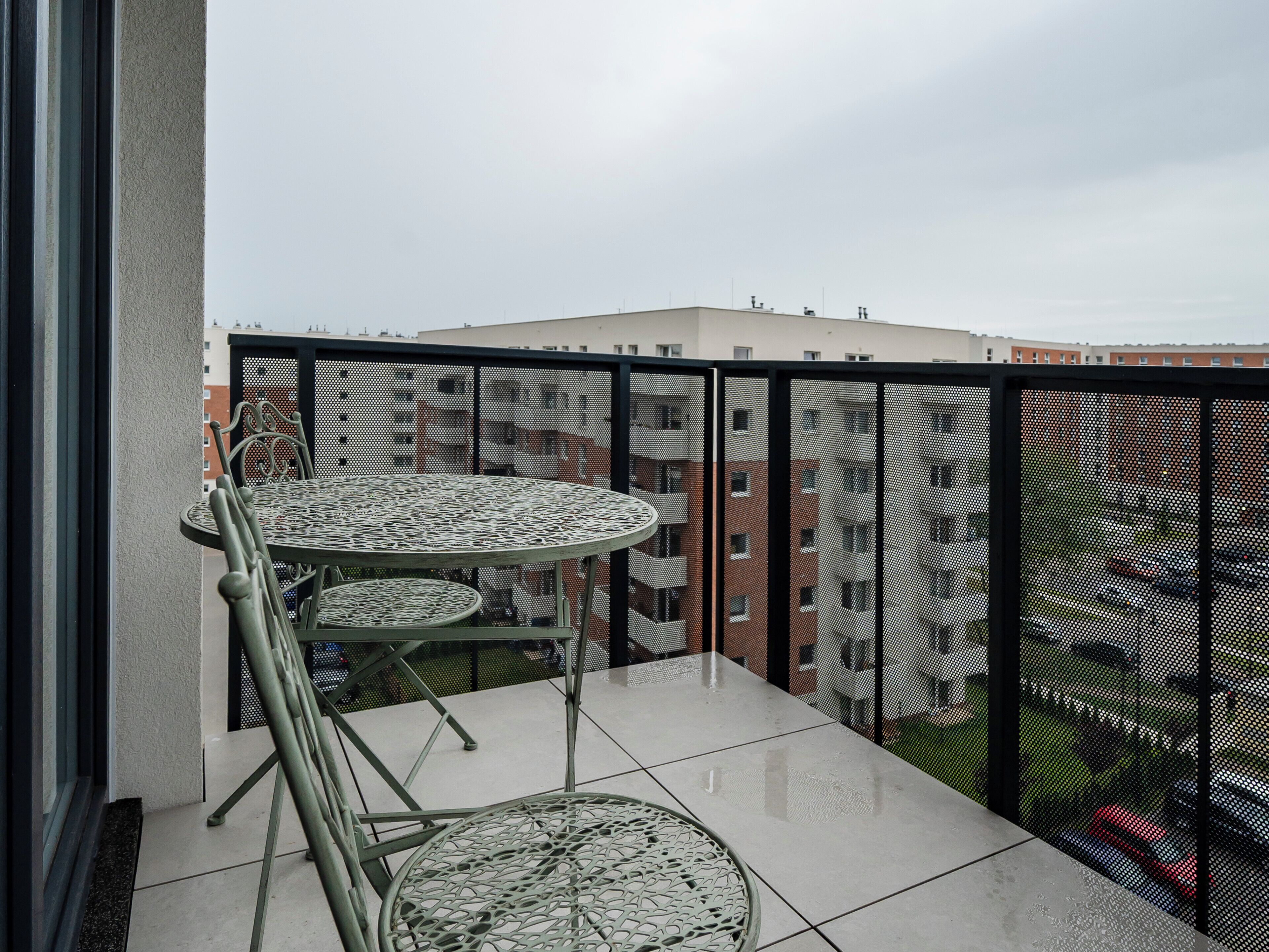 En balkon med et rundt bord og stole, gelænder i metal og udsigt til bygninger i baggrunden.