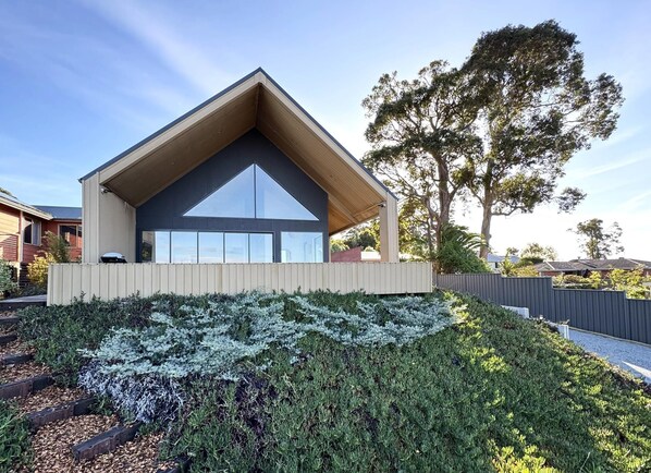 Une maison moderne dotée d’un toit triangulaire imposant, d’une grande fenêtre et d’un jardin bien entretenu où poussent différentes espèces de plantes.