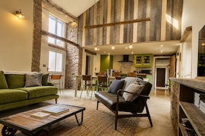 A spacious living area with a high ceiling, stone wall, and wooden beams. There is a green sofa, a leather armchair, and a coffee table with books. A dining area with chairs and a table is visible in the background. - Stonethwaite Barn, Dalston - a luxurious, dog friendly barn (Dalston)