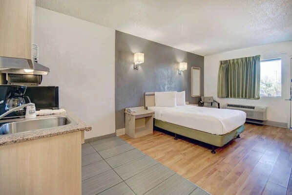 A hotel room with a bed, bedside table, a small kitchenette with a coffee maker, a window with curtains, and a wall-mounted heater. - Tucson Studio Getaway | Augie Acuna Los Ninos Park — 1 mile (Tucson)