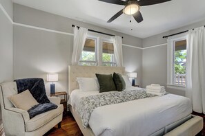 Ein Schlafzimmer mit einem Bett, einem Sessel, zwei Nachttischen, einem Deckenventilator und Fenstern mit Vorhängen.