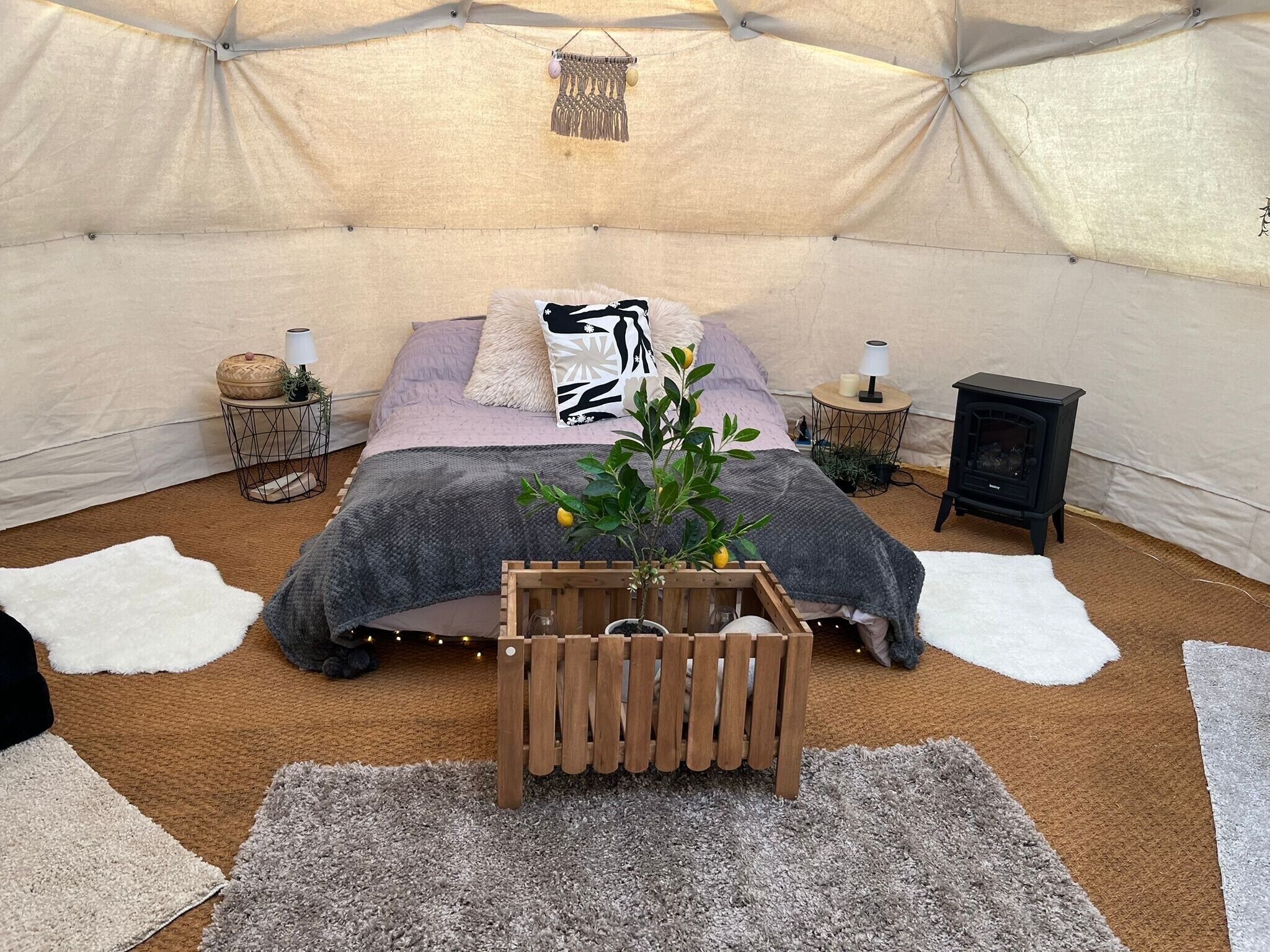 Een tent met een bed, nachtkastjes, een lamp, een kleine zwarte kachel en een houten kist met planten.