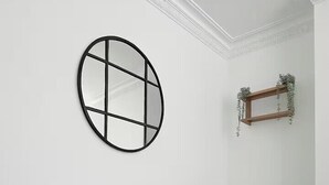Um sofá cinzento encostado a uma parede branca, com um espelho circular preto e uma prateleira de madeira com folhagem.