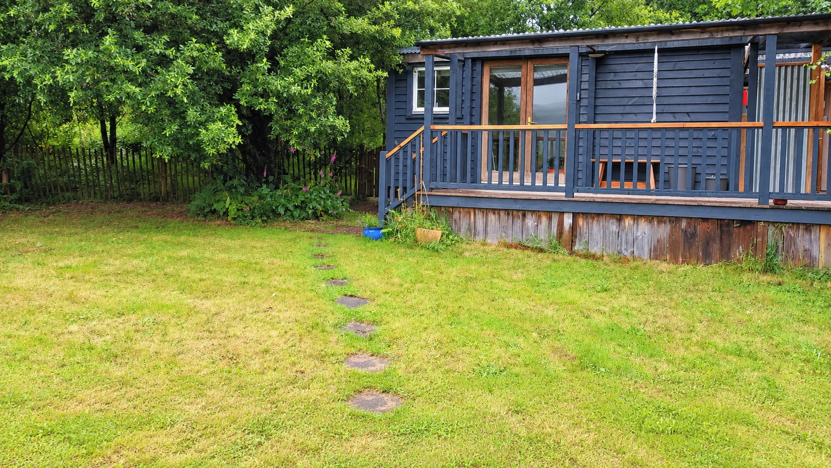 A wooden cabin with a deck, surrounded by a grassy yard and a bamboo fence.