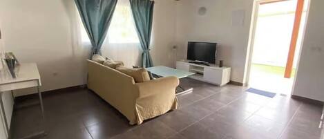 Una sala de estar con sofá, televisor sobre un mueble bajo, una mesita y una puerta que da al exterior.