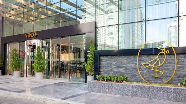 A modern hotel entrance with glass doors, a logo, and a stone wall with plants.