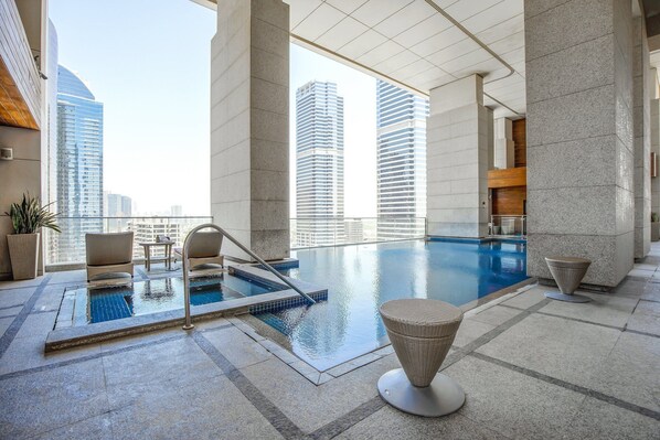 An indoor swimming pool with a view of tall buildings, stone columns, and wicker furniture.