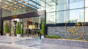 A modern hotel entrance with glass doors, a logo, and a stone wall with a bird design. - Deluxe Room | voco Bonnington | Pool, Sauna, Steam Rooms | 2 Units (Dubai)