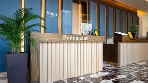 Un comptoir d’accueil avec un plateau en bois et un panneau vertical, une enseigne indiquant « Bonnington » et un logo « Voco » sur le mur.