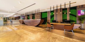 A modern reception area with a wooden reception desk, two chairs, a large potted plant, and a green wall with vertical wooden slats. - 1BR City | Mercure BH | Metro Facing, Pool, Squash Pool, Kids Club (Dubai)