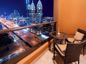 Un balcon doté d’une balustrade en verre, une table ronde avec deux chaises, et une vue sur le paysage urbain illuminé la nuit.