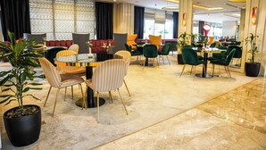 En moderne hotellobby med flere siddeområder, herunder runde borde og stole, samt en stor potteplante.