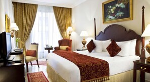 A hotel room with a large bed, two chairs, a desk with a television, and a painting on the wall.