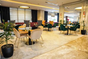 A modern hotel lobby with multiple seating areas, including round tables and chairs, and a large potted plant.