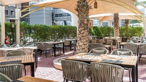 An outdoor cafe with tables and chairs, palm trees, and a modern building in the background.