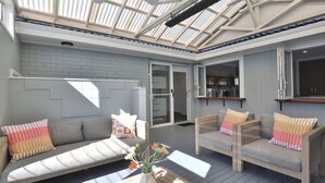 A spacious living area with a large glass door leading to a kitchen, a grey brick wall, and a wooden ceiling structure.