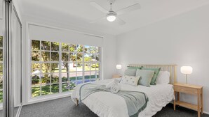 A bedroom with a bed, a nightstand, a lamp, a ceiling fan, and a large window with a view of a garden.