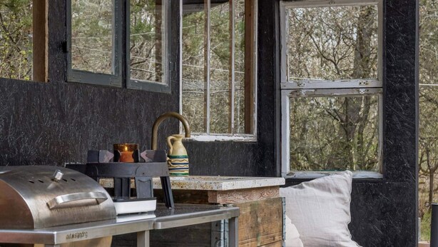 Ein rustikales Blockhaus-Interieur mit einem Metallgrill, Holzschränken und einem Fenster mit Blick auf Bäume.