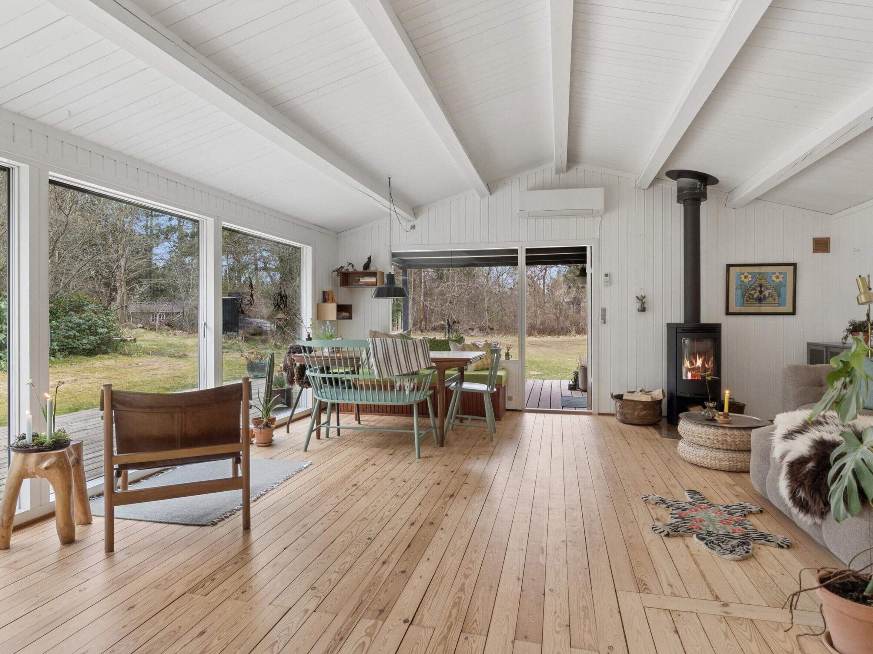 A spacious room with a wooden floor, a fireplace, a dining area with chairs, and a large window offering a view of the outdoors.