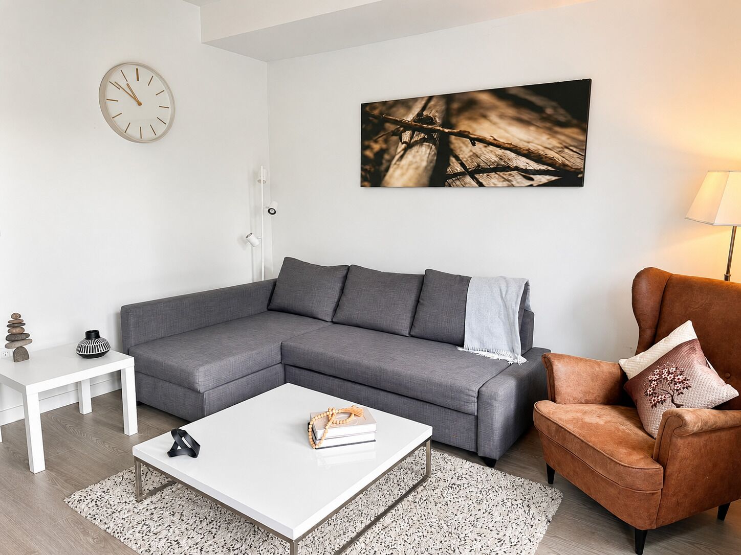 Ein modernes Wohnzimmer mit einem grauen Ecksofa, einem weißen Couchtisch, einem braunen Ledersessel, einem weißen Beistelltisch und einer Wanduhr.