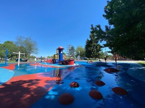 Ein Wasserpark mit einem Spielbereich in Blau und Rot, einer Wasserrutsche und einem großen Baum.