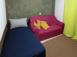 Una habitació petita amb un llit individual, un sofà vermell i un coixí groc.
