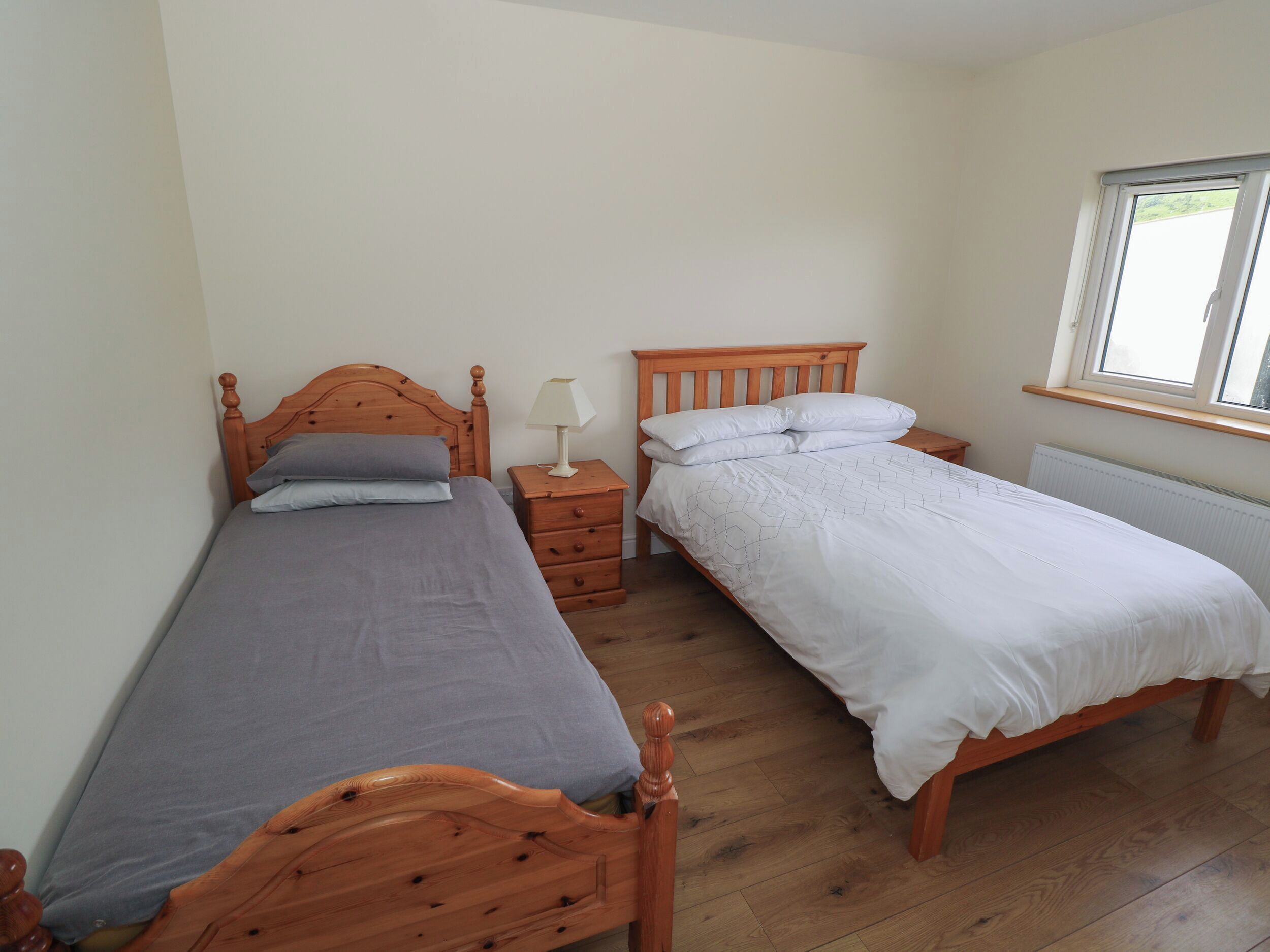 A room with two wooden beds, a nightstand with a lamp, a window, and a radiator.