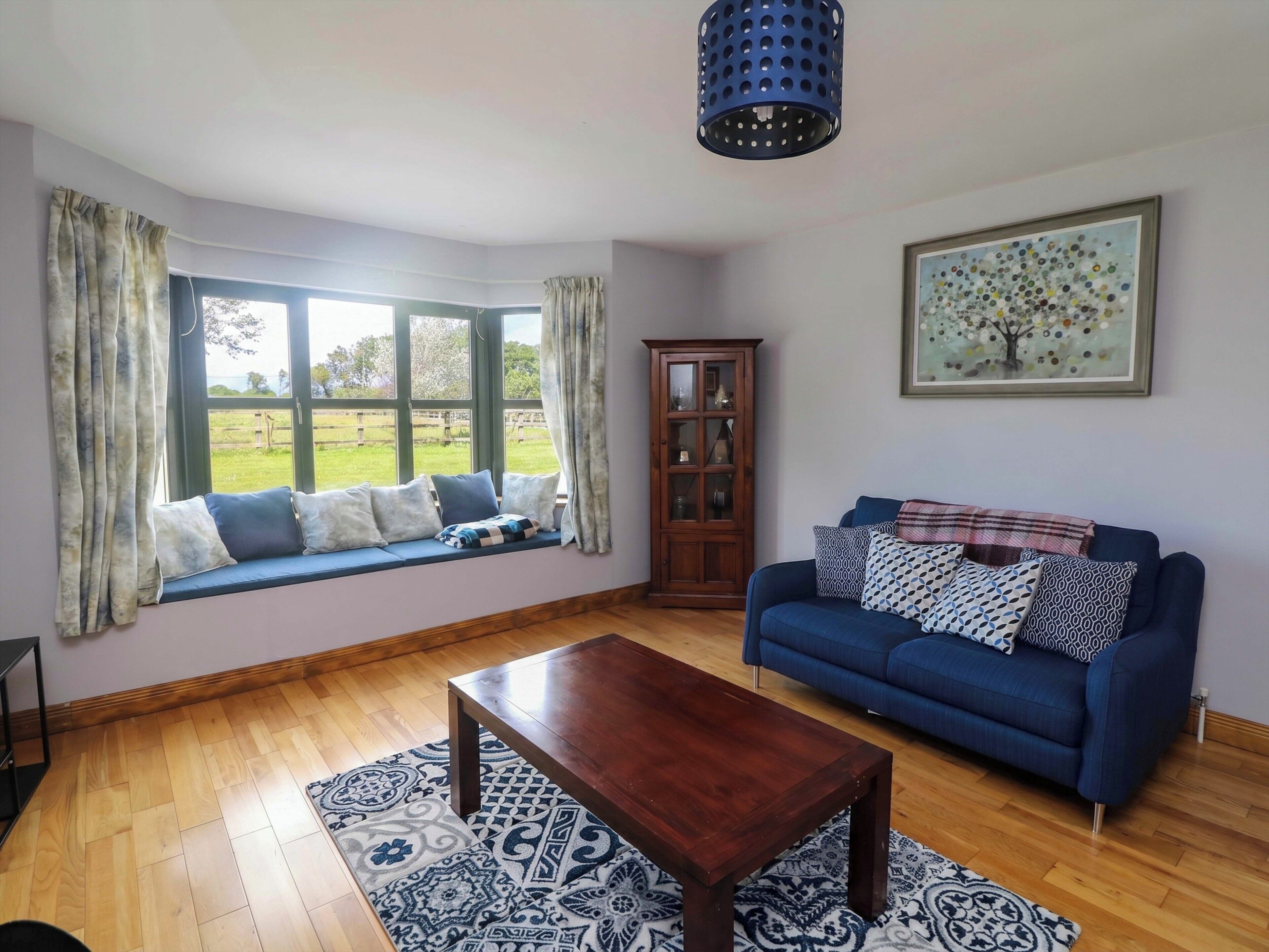 A living room with a blue sofa, a wooden coffee table, a window seat with cushions, a cabinet, and a painting on the wall.
