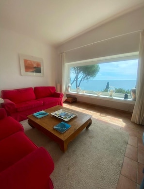 A living room with a red sofa, a wooden coffee table with magazines, a large window with a view of the sea, and a framed picture on the wall.