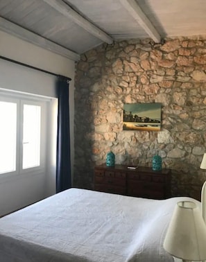 A bedroom with a stone wall, a bed, a dresser, and a window with curtains.