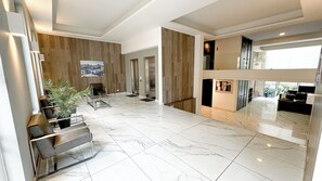Un vestíbulo espacioso con piso de mármol, un área de descanso con sillas y una mesa de centro, una pared decorativa de madera y vistas al exterior.