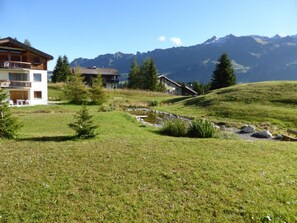 A hillside with a small stream, a wooden house, and a building with a balcony. - Cresta Sartons Brenn Appartement/Fewo, Badewanne, 1 Schlafraum by Interhome (Valbella)