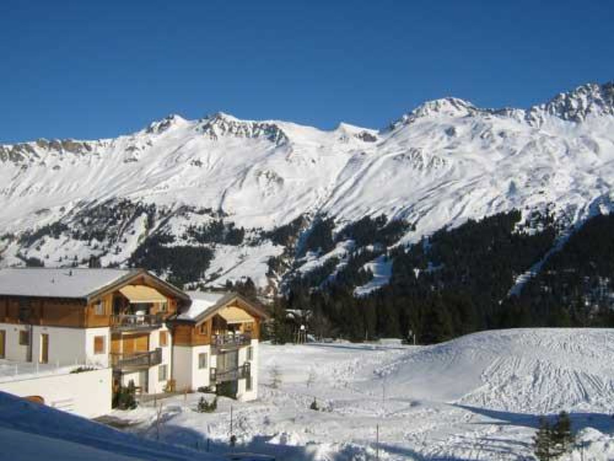 Een chalet met twee verdiepingen en een balkon, gelegen voor een achtergrond van besneeuwde bergen en een heldere lucht.