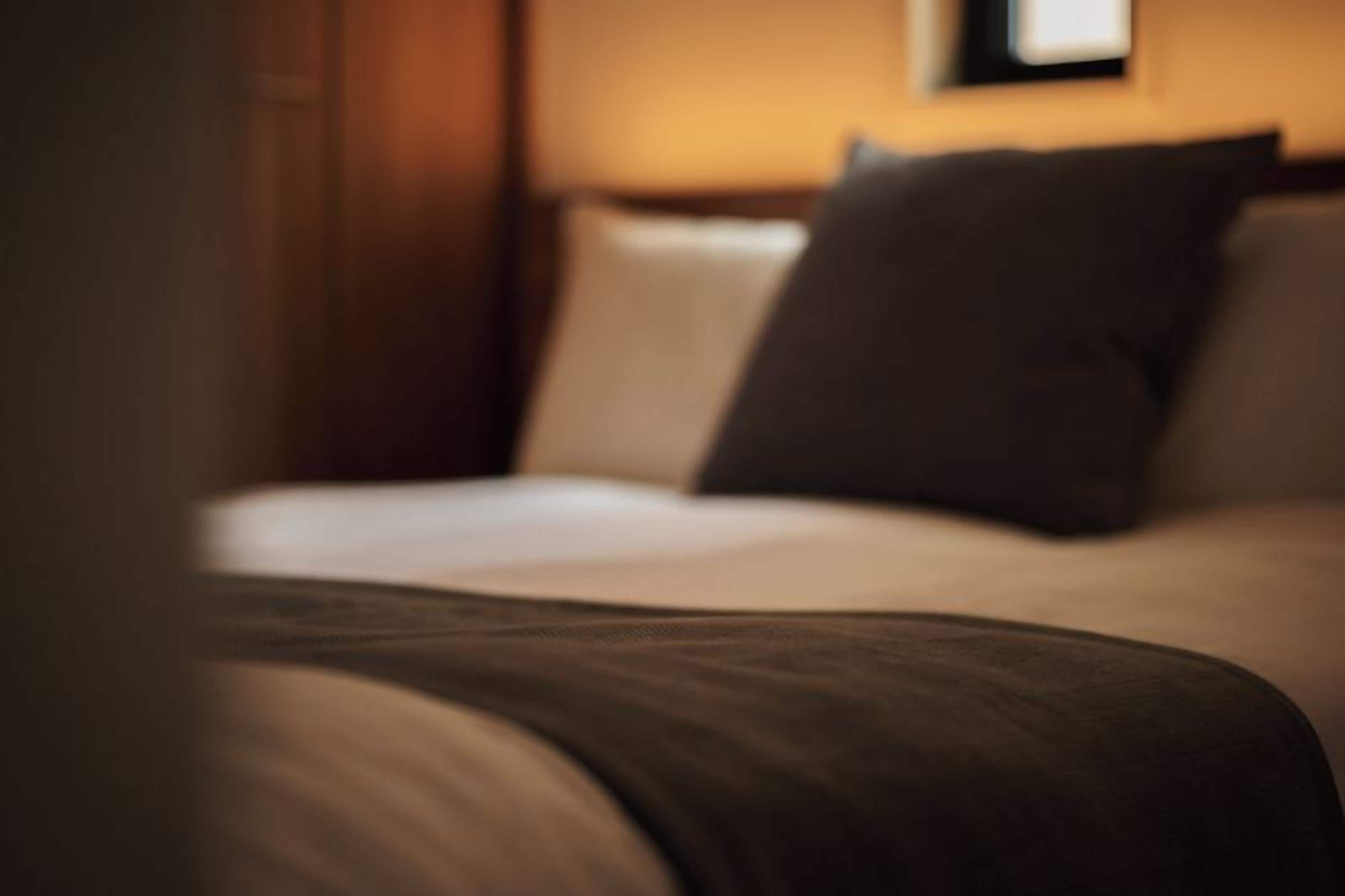 Una cama con sábana blanca y almohada oscura en una habitación con paredes de madera y un cuadro en la pared.