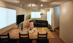 Uma sala de jantar com mesa de madeira, cadeiras e um vaso de flores. Há um sofá e uma televisão ao fundo.