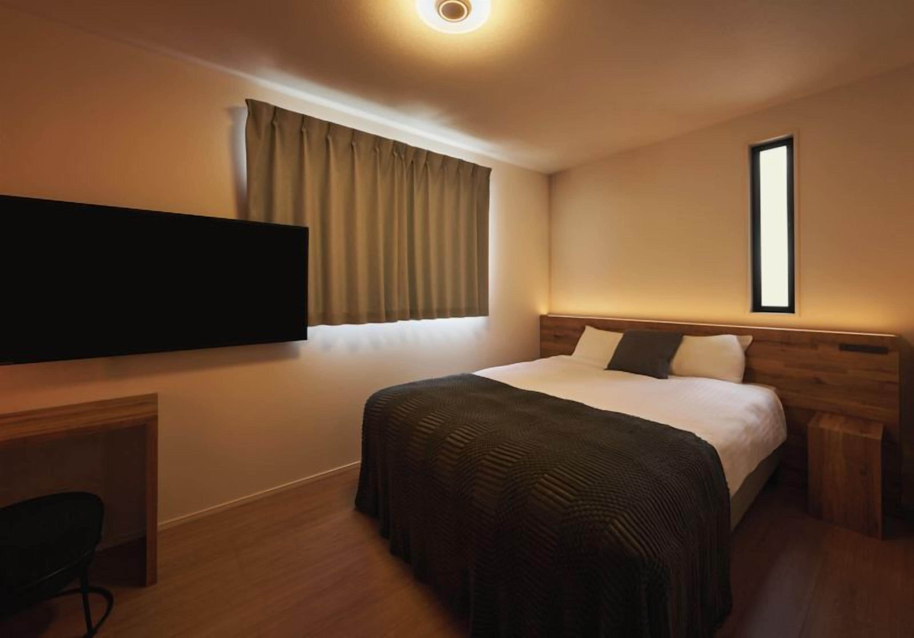 A hotel room with a bed, a television mounted on the wall, a desk, and a window with curtains.