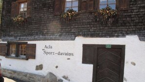 A building with a white exterior, wooden shutters, and a door marked with a green sign. - Acla Sporz Davains Appartement/Fewo, Dusche, WC, Wohn-/Schlafraum by Interhome (Lenzerheide)