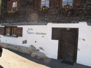 A building with a white exterior, wooden shutters, and a door marked with a green sign. - Acla Sporz Davains Appartement/Fewo, Dusche, WC, Wohn-/Schlafraum by Interhome (Lenzerheide)