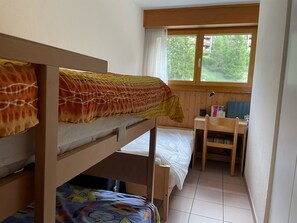 A dormitory room with bunk beds, a desk, a chair, and a window with a view of greenery outside.