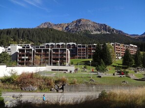 Un hotel de varios pisos con balcones, un estacionamiento y un paisaje montañoso al fondo.