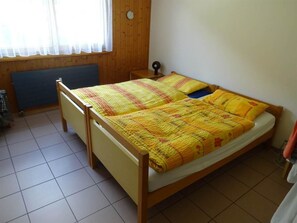 Ein Zimmer mit einem Einzelbett, Holzrahmen, einem Fenster mit durchscheinenden Vorhängen und einem Heizkörper unterhalb des Fensters.
