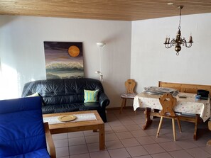 Un salon comprenant un canapé en cuir noir, une table basse en bois, une table à manger avec des chaises, un tableau représentant un paysage éclairé par la lune, une lampe sur pied et un lustre.