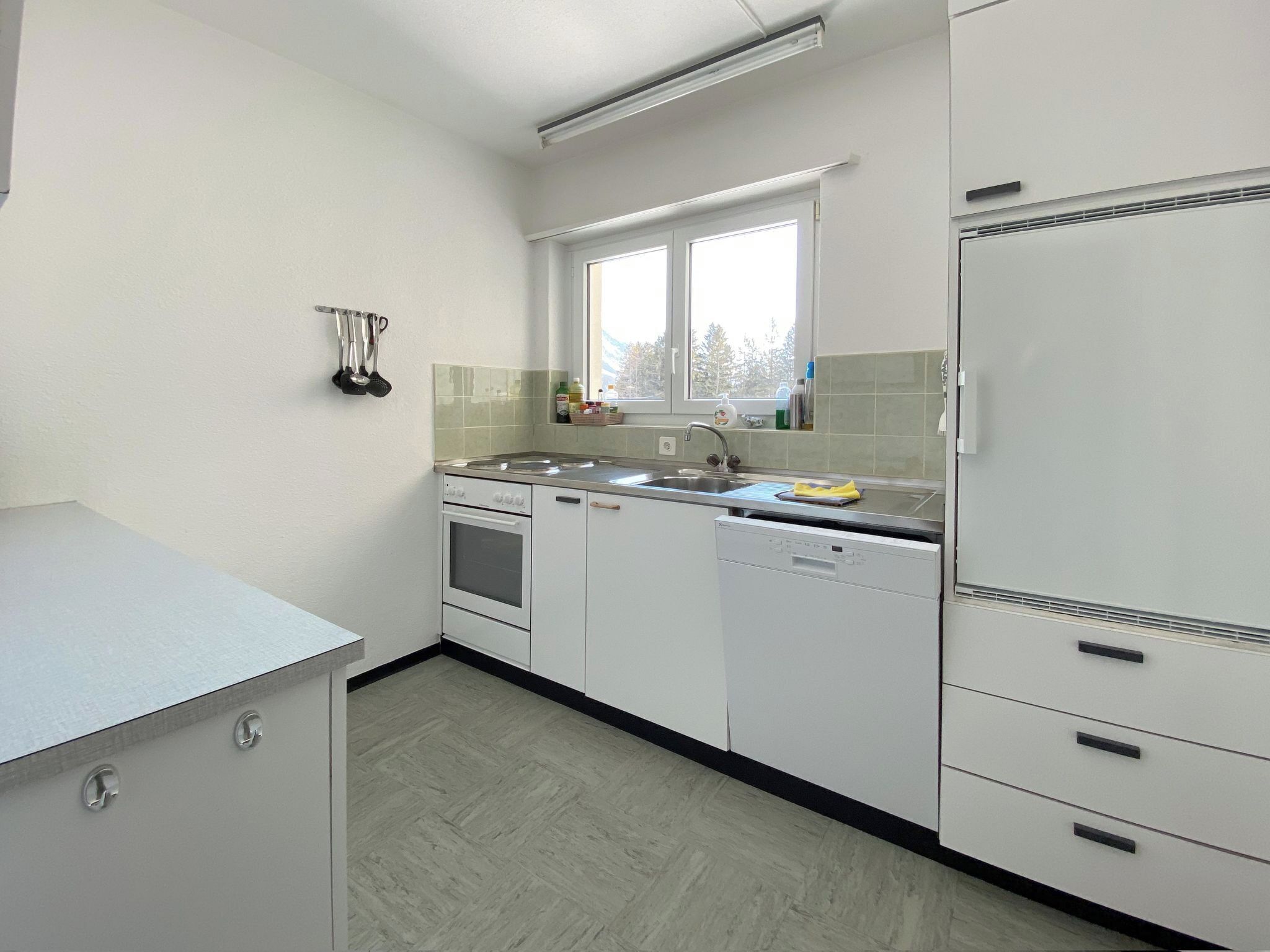 Una cucina con mobili bianchi, lavastoviglie, forno e una finestra vista alberi.