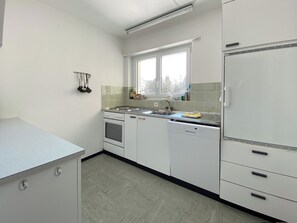 A kitchen with white cabinets, a dishwasher, an oven, and a window with a view of trees. - Haus Montana 1/9 Appartement/Fewo,   5-Bettwohnung by Interhome (Lenzerheide)
