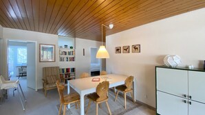 Un comedor con una mesa blanca, sillas de madera, una lámpara colgante y una estantería con libros.