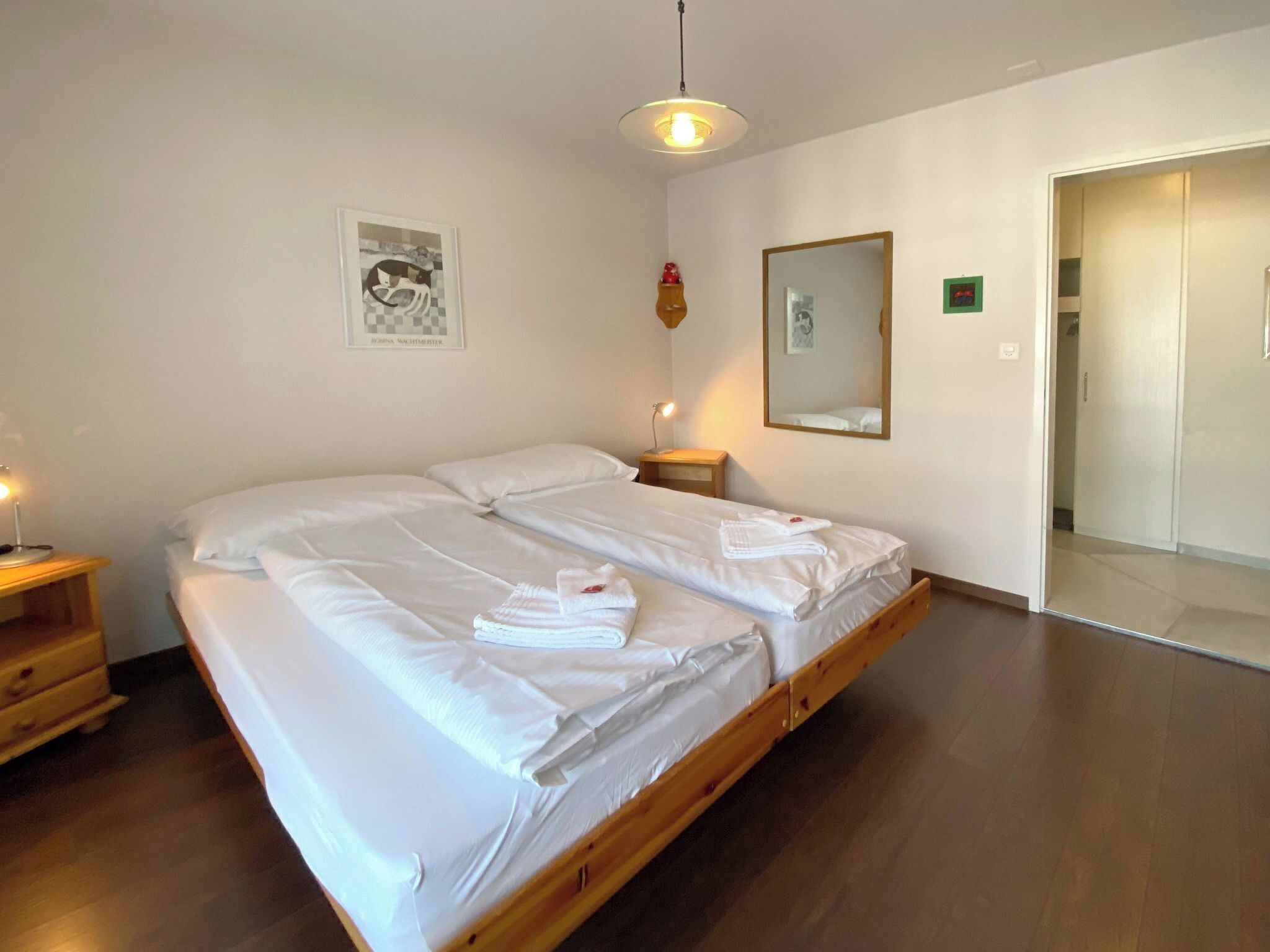A bedroom with a large bed, two bedside tables, a hanging light fixture, a mirror, and a door leading to another room.